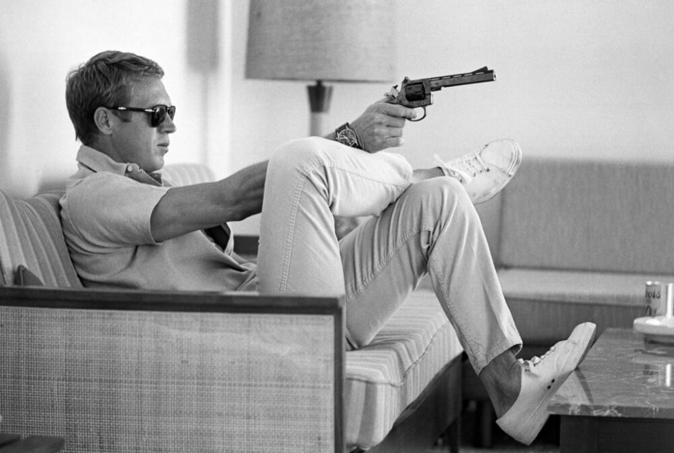 Profile view of American actor Steve McQueen (1930 - 1980) as he sits on a sofa in his home, sunglasses over his eyes, as he aims handgun, his wrist balanced on his crossed leg, Palm Springs, California, May 1963.